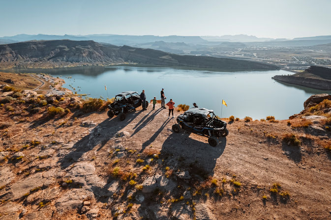Two off-road UTVs and a small group on a rocky desert overlook, enjoying a panoramic canyon and lake view in golden-hour light.