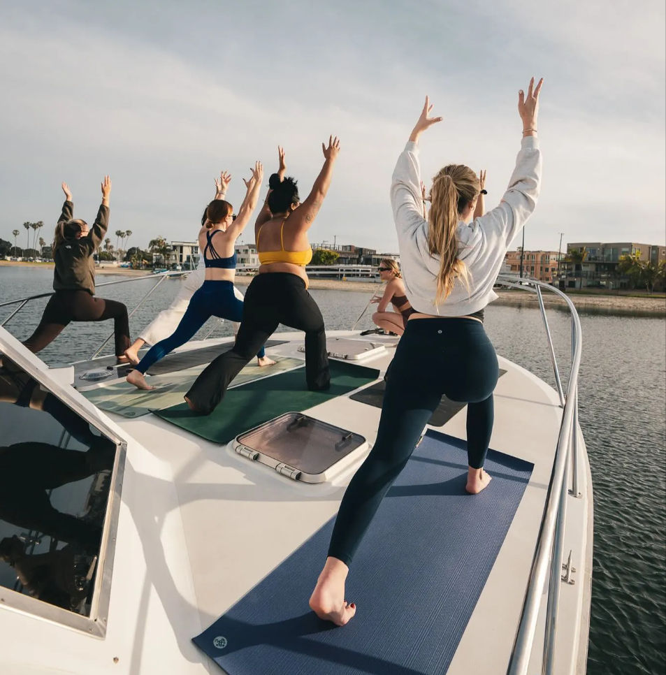 Group of people practicing yoga on mats on a boat deck at a sunny coastal marina, with waterfront buildings and palm trees in the background
