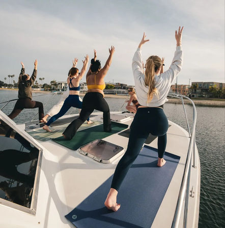 Group of people practicing yoga on mats on a boat deck at a sunny coastal marina, with waterfront buildings and palm trees in the background