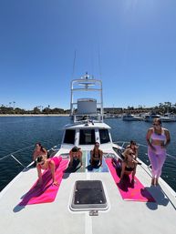 Sun-soaked yoga session: women on colorful mats practicing on the bow of a white yacht in a palm-lined coastal marina under a clear blue sky.