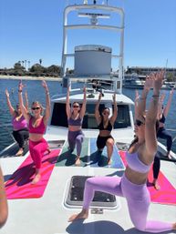 Group of women practicing yacht yoga on colorful mats atop a white boat at a sunny marina, with palm-lined waterfront and moored boats in the background.