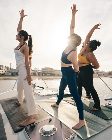 Three women practicing boat yoga on mats aboard a yacht at sunset, reaching arms skyward with sun flare and a calm marina with waterfront homes in the background.