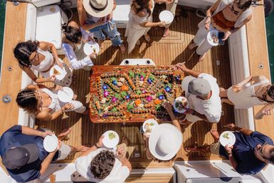 Aerial view of people on a wooden yacht deck sharing a giant seafood platter decorated with colorful edible flowers — summer boat party on turquoise water.