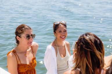 Three friends laughing on a sunny boat trip — a woman in a rust sundress with sunglasses, another in a white cover-up holding a wine glass, and a third seen from behind with sparkling blue water behind them.