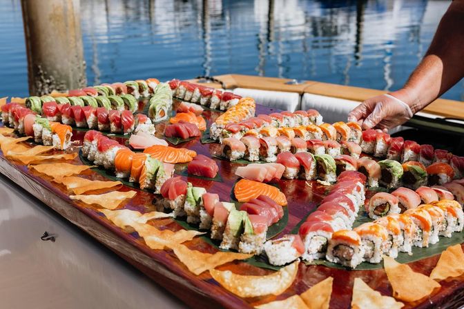 Vibrant assorted sushi platter on a large wooden board dockside — rows of tuna, salmon and avocado-topped rolls, sashimi slices and crispy wonton chips with a gloved hand arranging pieces.
