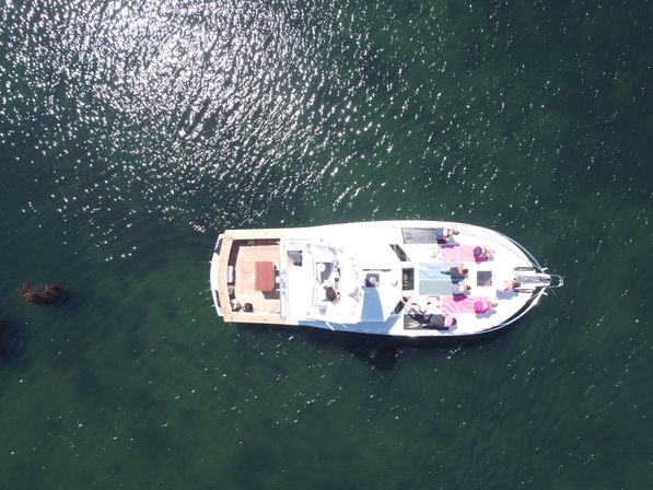 Aerial drone view of a white yacht floating in green water with sunlight sparkling on the surface, passengers sunbathing on colorful towels at the bow and a wooden aft deck visible.