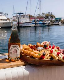 Sparkling rosé bottle beside a wooden serving board of croissants, mini waffles, pancakes and fresh berries on a boat railing with sunlit sailboats and a calm marina harbor in the background