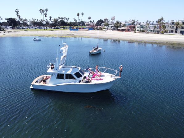 White motor yacht anchored in a calm blue bay near a sandy beach lined with palm trees; a group sunbathing and lounging on the bow while one person stands at the front taking photos.