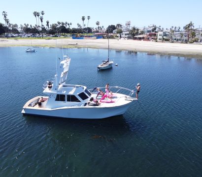 White motor yacht anchored in a calm blue bay near a sandy beach lined with palm trees; a group sunbathing and lounging on the bow while one person stands at the front taking photos.