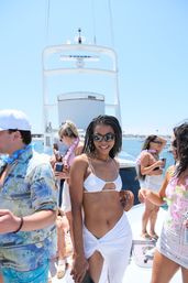 Smiling woman in a white bikini and sarong wearing sunglasses at a sunny yacht party, friends in tropical shirts and leis on the deck with ocean and marina in the background.
