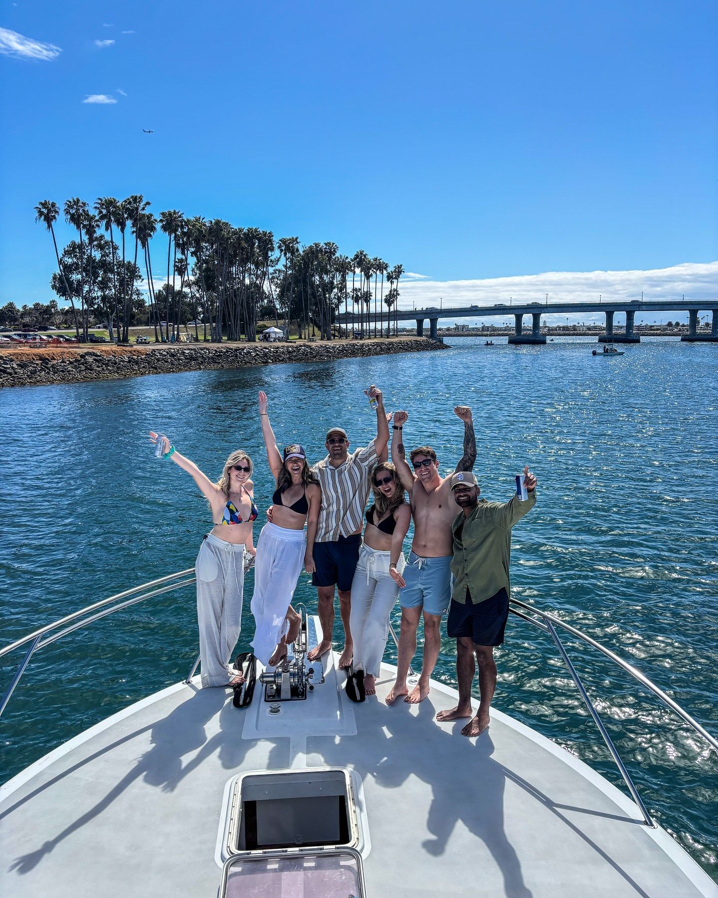 Six people cheering on the bow of a yacht during a sunny bay cruise, sparkling blue water, palm trees along the shoreline and a low bridge in the distance.