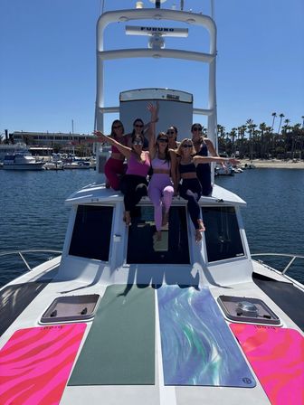 Seven women in colorful athleisure posing and laughing on the bow of a white yacht at a sunny coastal marina with palm trees and docked boats.