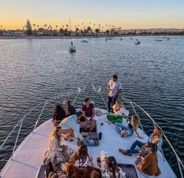 Group of people relaxing on the bow of a yacht at sunset in a palm-lined harbor with anchored sailboats and a coastal skyline