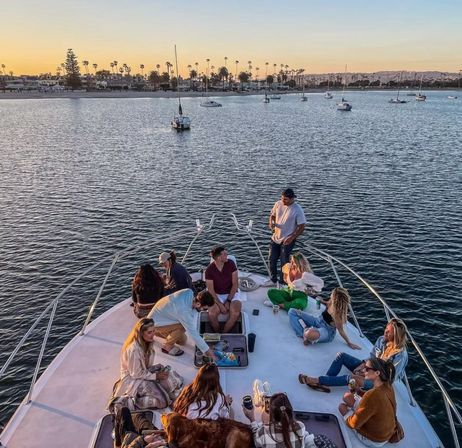 Group of people relaxing on the bow of a yacht at sunset in a palm-lined harbor with anchored sailboats and a coastal skyline