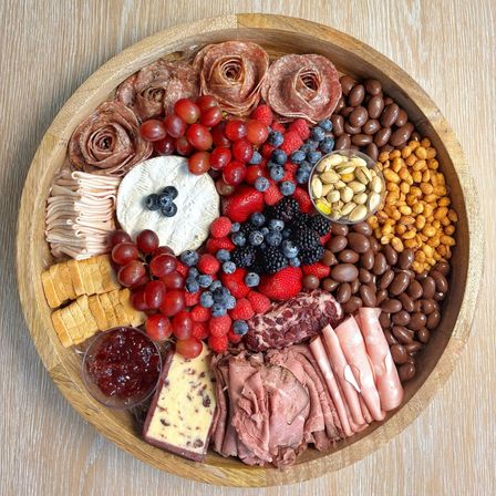 Round wooden charcuterie board with a brie wheel, salami roses, roast beef and folded ham, cranberry-studded cheese, crackers, grapes, strawberries, blueberries, raspberries, blackberries, pistachios, chocolate-covered nuts, crunchy corn nuts and red jam.