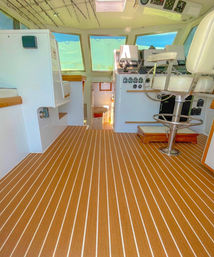 Bright yacht helm interior with striped teak-style flooring, white pedestal captain chair, instrument console, overhead rod storage and steps down to cabin with ocean view