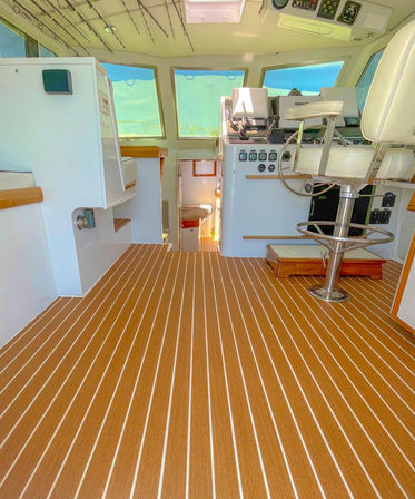 Sunlit yacht helm cabin with teak-style striped marine flooring, white captain's chair and navigation console, panoramic windows showing turquoise sea and steps down to a cozy lower berth.