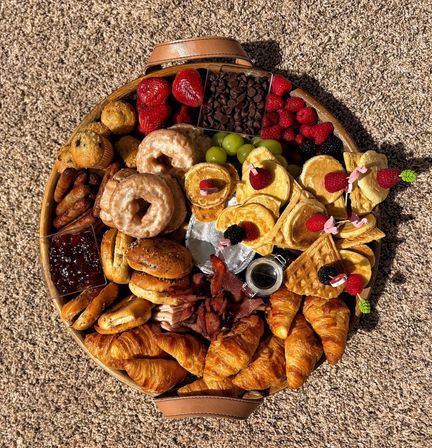 Round breakfast grazing board on carpet with buttery croissants, glazed donuts, mini pancakes and waffles topped with berries, bagels and muffins, bacon, grapes, chocolate chips and jam — colorful brunch platter