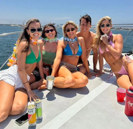 Five friends in colorful bikinis and leis enjoying a sunny boat party, smiling with drinks on deck against a blue ocean and coastline backdrop.
