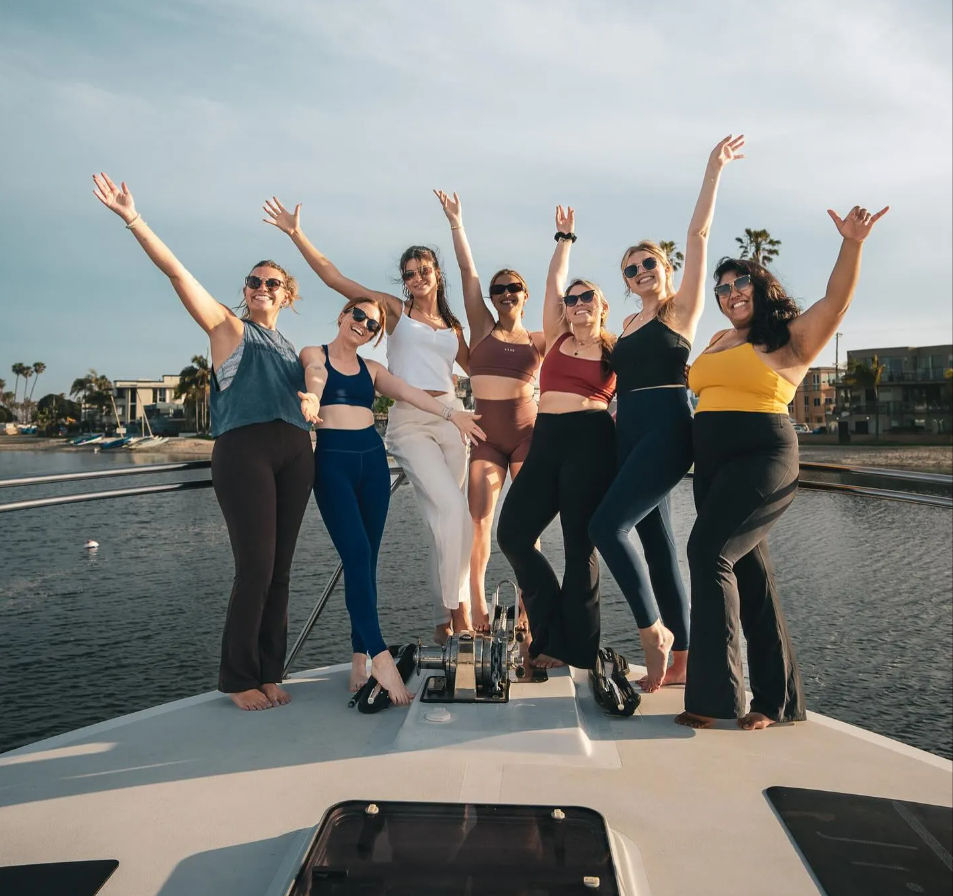 Seven women in activewear and swimsuits cheering with arms raised on the bow of a yacht at a sunny coastal marina with palm trees and waterfront homes