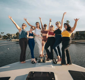 Seven women in activewear and swimsuits cheering with arms raised on the bow of a yacht at a sunny coastal marina with palm trees and waterfront homes