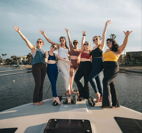 Seven women in activewear and swimsuits cheering with arms raised on the bow of a yacht at a sunny coastal marina with palm trees and waterfront homes