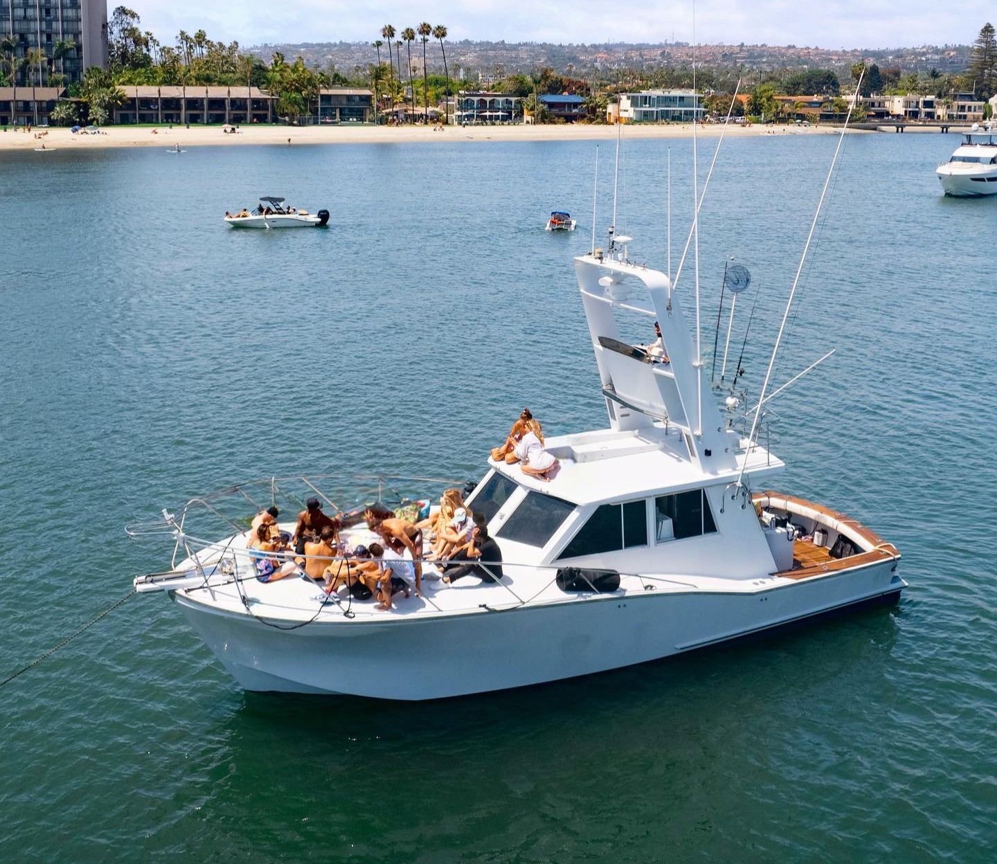 White motor yacht anchored in a calm coastal bay with a group of people lounging and sunbathing on the bow, palm-lined sandy beach and waterfront buildings in the background on a sunny day — lively boat party scene.