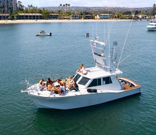 White motor yacht anchored in a calm coastal bay with a group of people lounging and sunbathing on the bow, palm-lined sandy beach and waterfront buildings in the background on a sunny day — lively boat party scene.