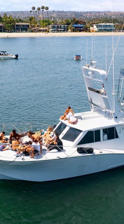 White motor yacht anchored in a calm coastal bay with a group of people lounging and sunbathing on the bow, palm-lined sandy beach and waterfront buildings in the background on a sunny day — lively boat party scene.