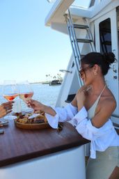 Two people clinking rosé wine glasses with strawberries aboard a yacht, wooden snack board on the table and a sunny waterfront marina with palm trees in the background.