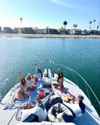Group of friends relaxing and waving on the bow of a white yacht in turquoise coastal waters on a sunny day, with a sandy beach and palm-lined shore in the background.