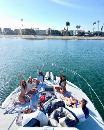 Group of friends relaxing and waving on the bow of a white yacht in turquoise coastal waters on a sunny day, with a sandy beach and palm-lined shore in the background.