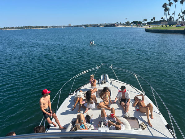 Friends lounging and sunbathing on the bow of a white yacht in a sunny coastal bay with palm trees and calm blue water.