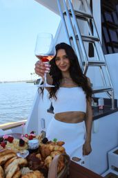 Person on a yacht toasting with a glass of rosé over a brunch spread of croissants and pastries, sunny coastal bay and palm trees in the background.