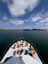 Group of people relaxing and sunbathing on the bow of a white yacht cruising a calm coastal bay near a marina, blue water and scattered clouds overhead — summer boat outing.