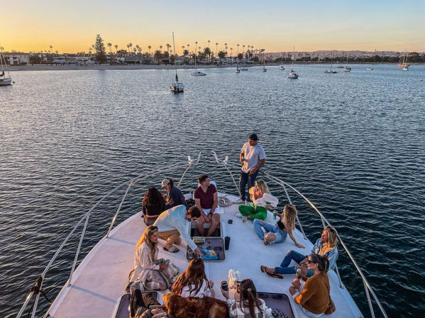 Group of friends relaxing on the bow of a yacht at sunset, anchored in a calm coastal marina with sailboats and a palm‑tree lined shoreline.
