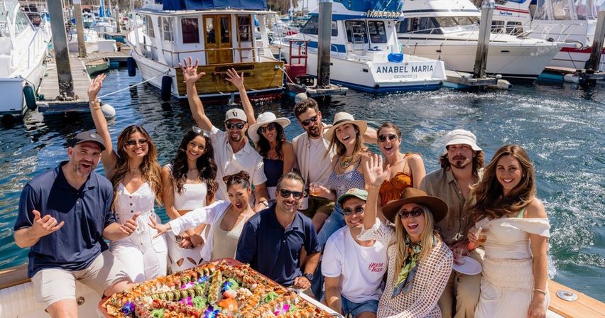 Cheerful group of friends on a sunny boat at a marina, waving and posing around a large colorful seafood and sushi platter with yachts and docks in the background.