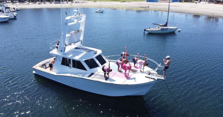 White motor yacht anchored in a calm coastal bay with a group in pink workout clothes doing yoga poses on the bow while a photographer snaps pictures; sailboats and a sandy beach sit in the background.
