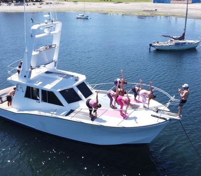 White motor yacht anchored in a calm coastal bay with a group in pink workout clothes doing yoga poses on the bow while a photographer snaps pictures; sailboats and a sandy beach sit in the background.
