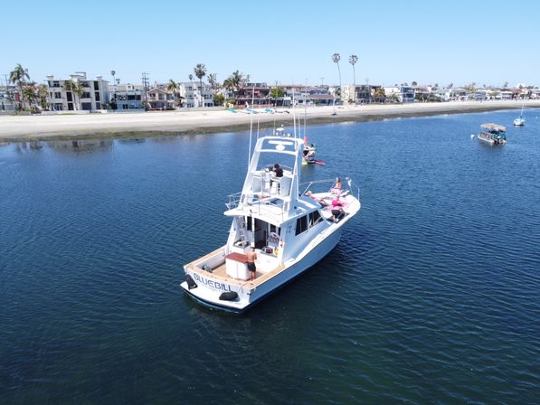 White motor yacht anchored in a calm blue bay near a sandy beachfront lined with palm trees and homes, with people relaxing on deck and small boats and kayaks nearby.