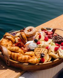 Sunny waterfront breakfast charcuterie board on a wooden table with croissants, mini pancakes and waffles, donuts, bagels, bacon, fresh berries and chocolate chips