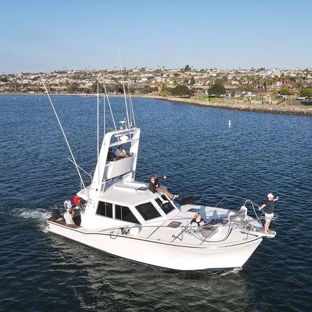 White sportfishing yacht cruising in a calm coastal bay near a shoreline with seaside homes, people relaxing on deck under a clear sunny sky.