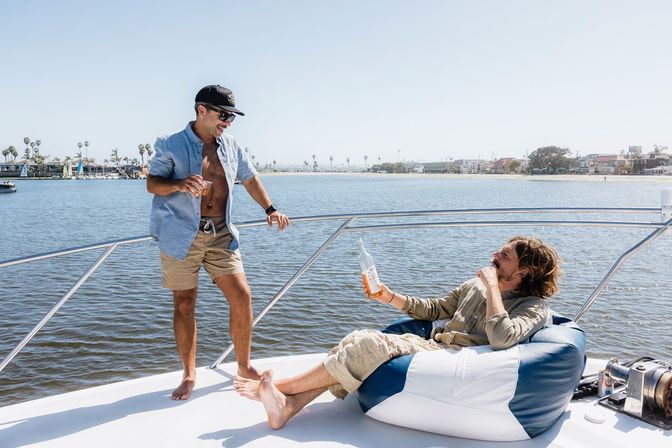 Two friends relaxing on a sunny yacht in a coastal bay — one standing with a drink, the other lounging on a blue-and-white bean bag holding a bottle, palm-lined marina in the background.