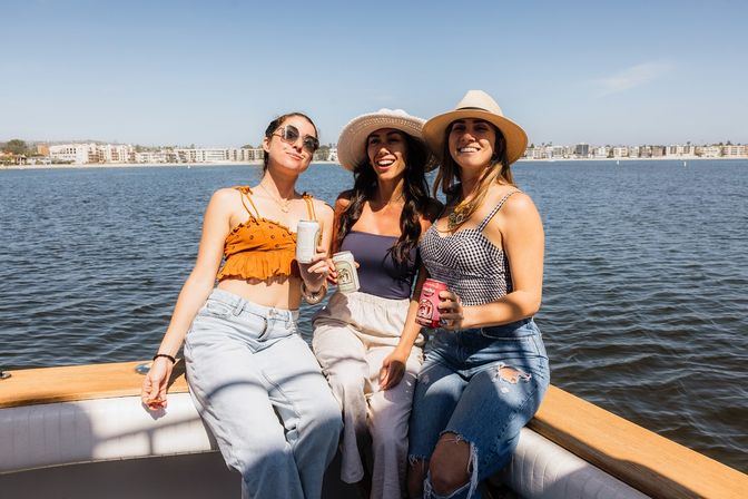 Three friends laughing on a sunny boat ride in a coastal bay, wearing summer hats and casual outfits while holding canned drinks with waterfront buildings in the distance.