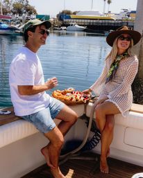 Smiling friends on a boat at a sunny coastal marina sharing a pastry brunch platter, wearing sun hats, sunglasses and casual summer clothes