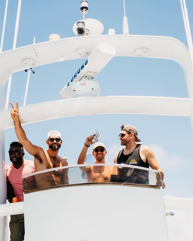 Four friends celebrating on the upper deck of a white yacht under a clear blue sky, wearing caps and sunglasses, one holding a drink and another flashing a peace sign.