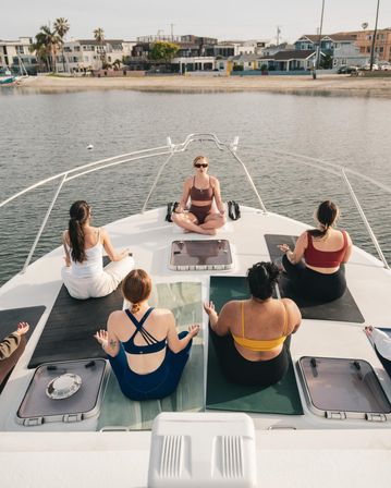 Sun-soaked group yoga on the bow of a boat in a calm coastal bay, instructor seated at the front, participants on mats facing the water and beachfront homes.