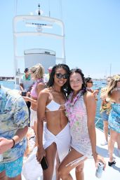 Two women smiling on a sunny yacht deck in swimwear and floral leis, wearing sunglasses and holding drinks at a lively boat party near a marina