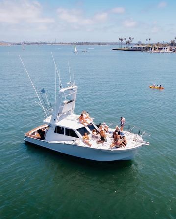 White motor yacht anchored in a sunny coastal bay with people socializing and sunbathing on deck, sailboats and a palm-lined marina on the horizon and a kayaker nearby.