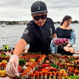 Sushi chef arranging a vibrant sushi spread on a wooden board at a waterfront event — rows of colorful rolls, sashimi, seaweed salad and orchid garnishes.